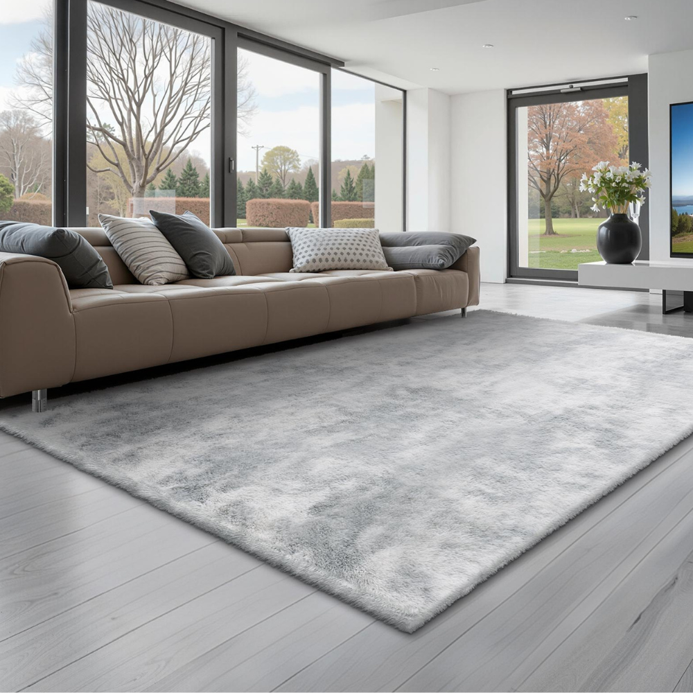 Contemporary living room with a large, gray area rug on light wood flooring, a tan sofa with assorted pillows, and floor-to-ceiling windows offering a view of a landscaped garden.