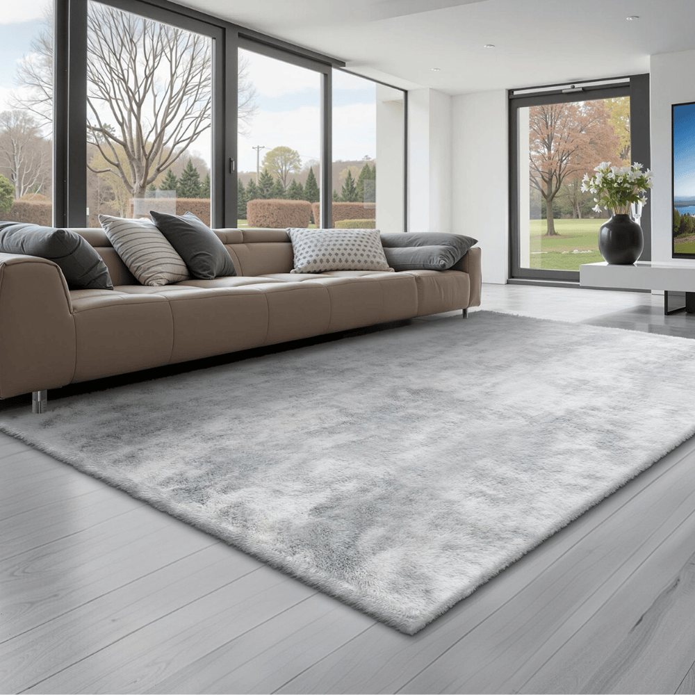Contemporary living room with a large, gray area rug on light wood flooring, a tan sofa with assorted pillows, and floor-to-ceiling windows offering a view of a landscaped garden.