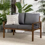 Wooden loveseat with dark gray cushions and a decorative pillow, surrounded by various potted plants against a white shiplap wall.