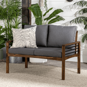 Wooden loveseat with dark gray cushions and a decorative pillow, surrounded by various potted plants against a white shiplap wall.
