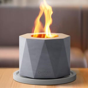 A geometric tabletop fire pit with a hexagonal design and a visible flame, made from gray stone-like material, resting on a matching circular base. It features a \“CAUTION HOT\“ warning on the side.