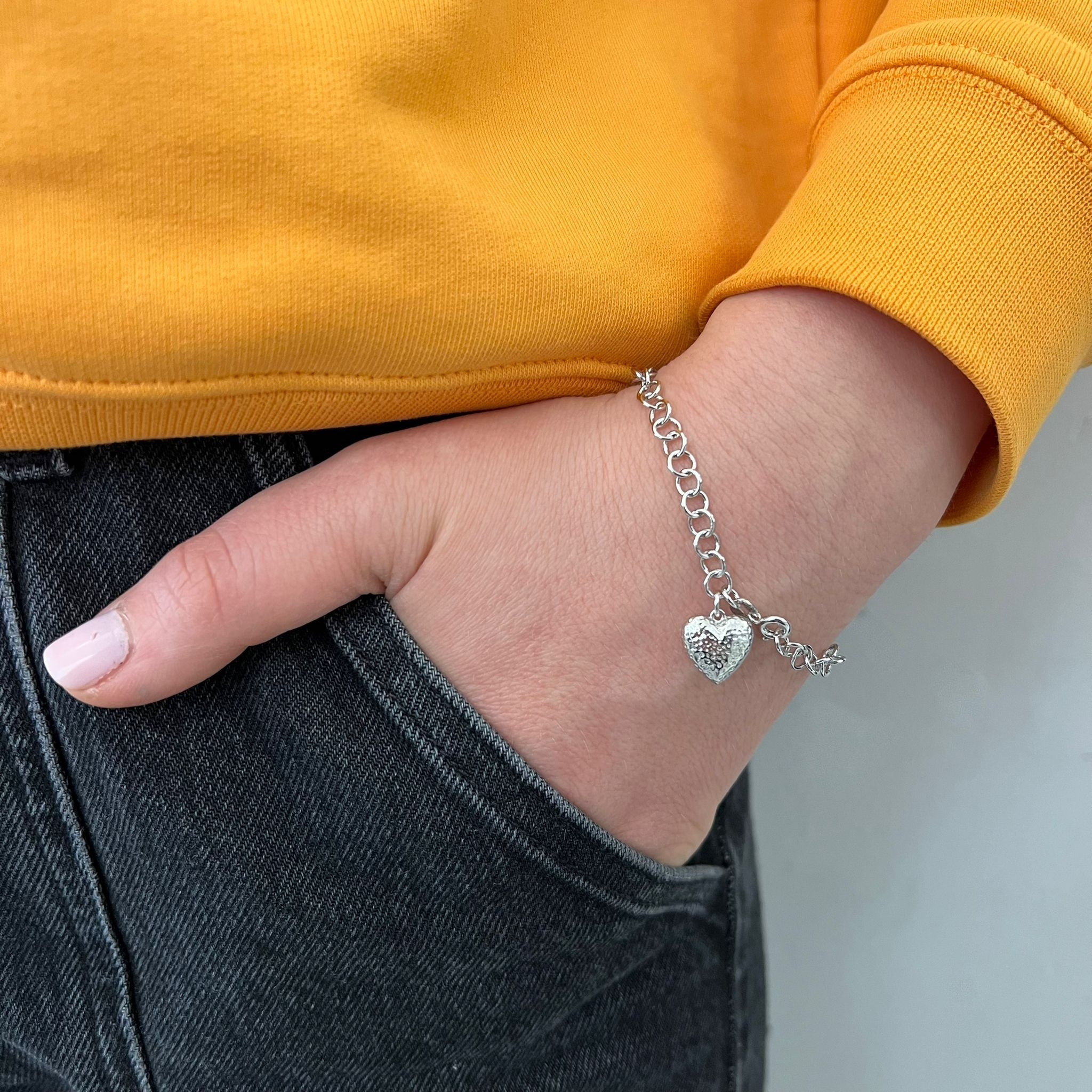 A silver bracelet with a heart charm, worn on a wrist against a yellow top and gray pants.