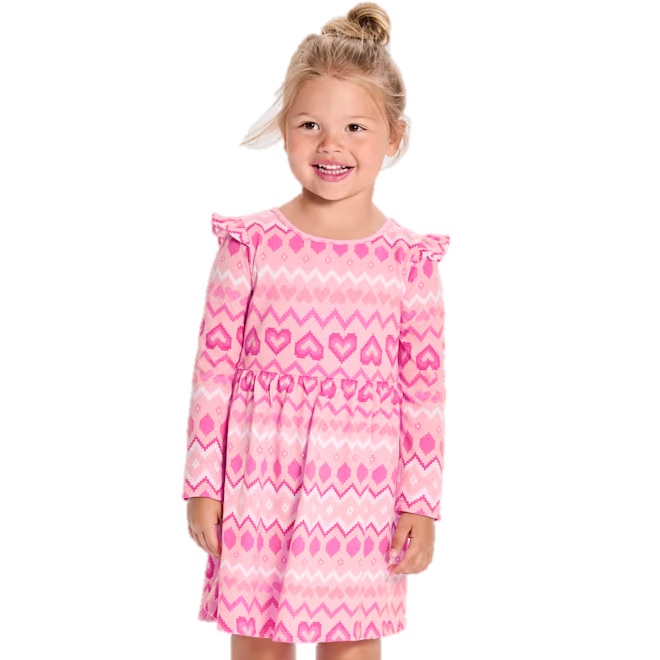 A child is wearing a pink long-sleeve dress featuring a pattern with hearts and zigzags. The dress has ruffled shoulders.