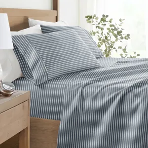 A neatly made bed features a blue and white striped sheet set with matching pillowcases. A wooden side table holds a striped lamp, a small plant, and a clock.