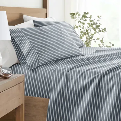 A neatly made bed features a blue and white striped sheet set with matching pillowcases. A wooden side table holds a striped lamp, a small plant, and a clock.