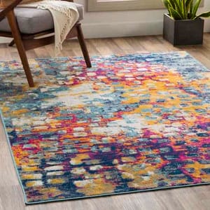A colorful abstract area rug with a mix of vibrant blues, yellows, pinks, and whites, measuring 5' x 7', displayed on a light wooden floor next to a modern chair and a potted plant.