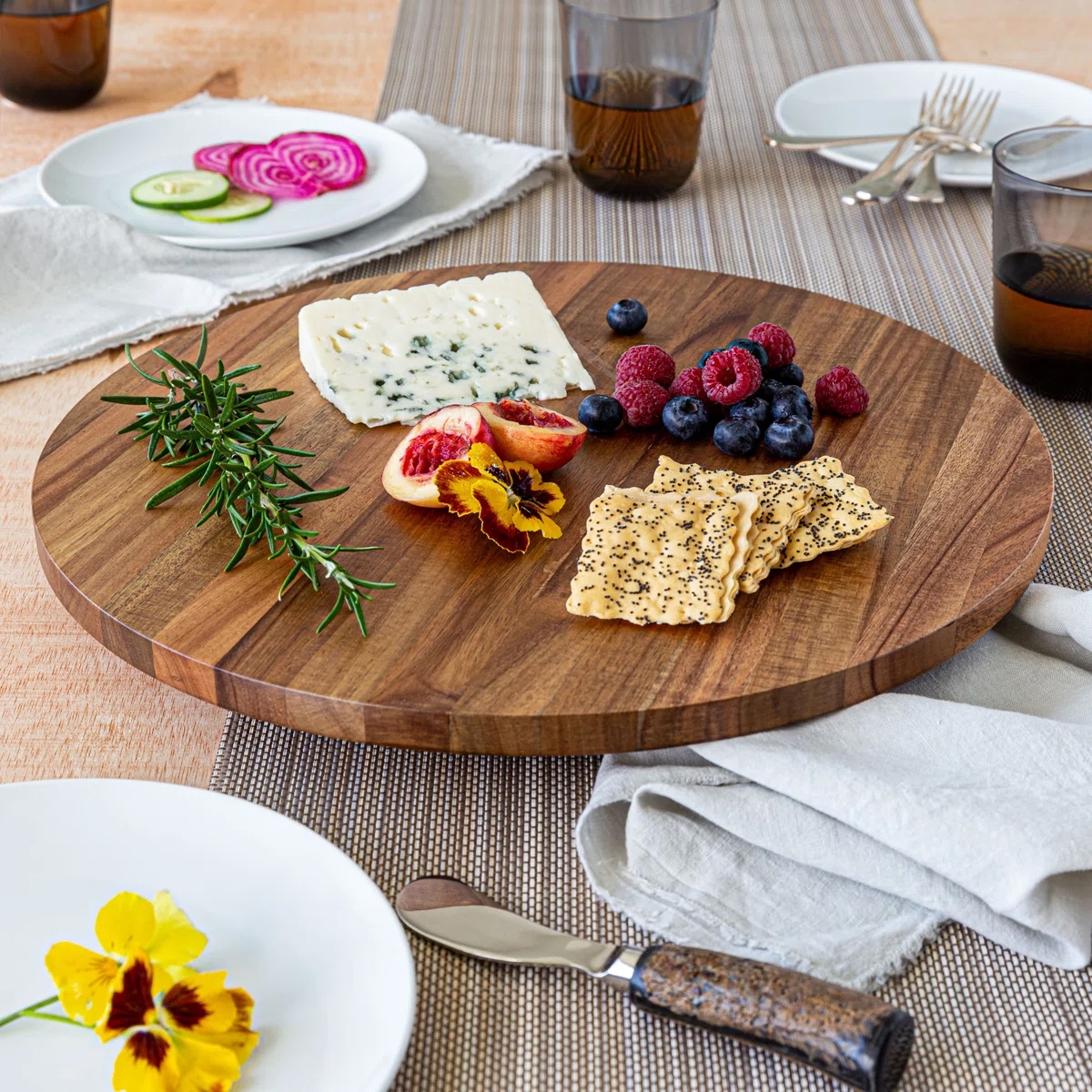 A round wooden serving board with cheese, crackers, rosemary, blueberries, raspberries, blood orange slices, and edible flowers, set on a table with plates and glasses.