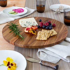 A round wooden serving board with cheese, crackers, rosemary, blueberries, raspberries, blood orange slices, and edible flowers, set on a table with plates and glasses.