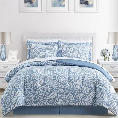 A bedding set featuring a light blue and white floral pattern, including a comforter, two pillow shams, a flat sheet, a fitted sheet, two pillowcases, and a bed skirt. The room decor includes lamps, floral arrangements, and abstract wall art.