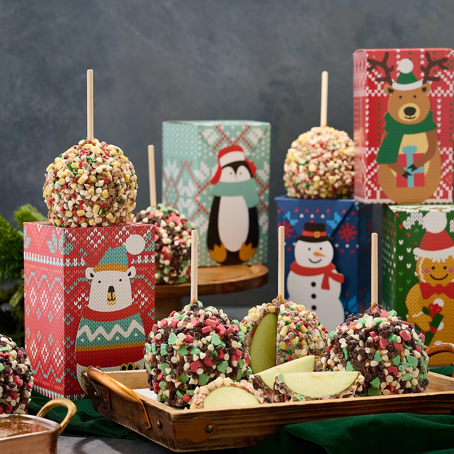 Candy apples coated in colorful sprinkles are displayed with festive patterned boxes featuring holiday characters in the background.