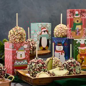 Candy apples coated in colorful sprinkles are displayed with festive patterned boxes featuring holiday characters in the background.