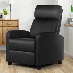 A black recliner chair with a sleek design is positioned on a wooden floor, surrounded by light-colored furniture and a few decorative elements like plants and stacked books.