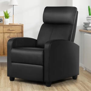 A black recliner chair with a sleek design is positioned on a wooden floor, surrounded by light-colored furniture and a few decorative elements like plants and stacked books.