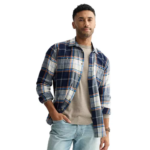 A man wears a blue and white plaid shirt over a beige sweater with light blue jeans.