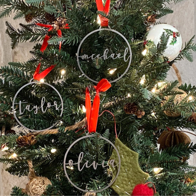 Personalized Christmas tree ornaments with names \“Taylor,\“ \“Michael,\“ and \“Olivia\“ written in script font, hung with red ribbons on a decorated tree with lights and other ornaments.