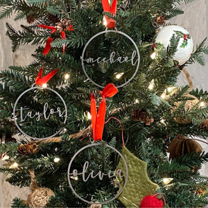 Personalized Christmas tree ornaments with names \“Taylor,\“ \“Michael,\“ and \“Olivia\“ written in script font, hung with red ribbons on a decorated tree with lights and other ornaments.