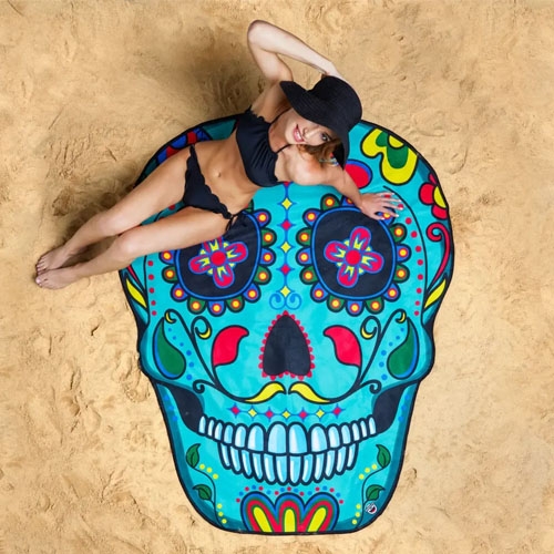 A vibrant beach blanket shaped like a sugar skull with colorful, intricate floral and geometric designs, placed on sand. A person lounges on it, highlighting its size and vivid patterns.