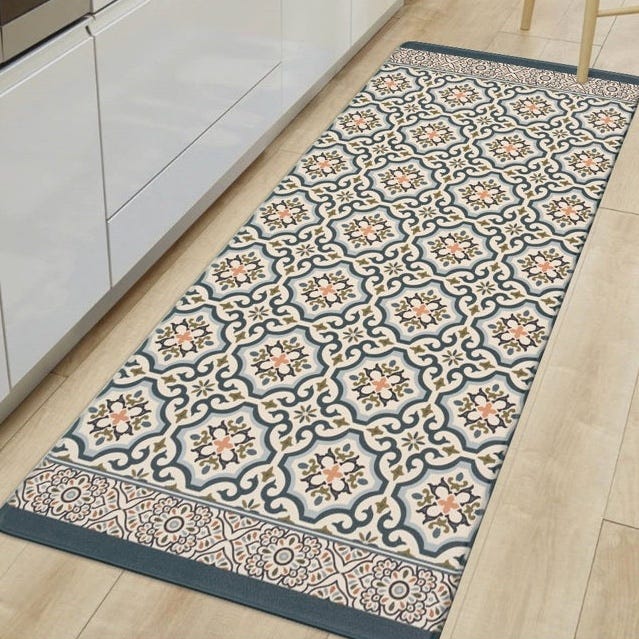 A rectangular kitchen runner rug with a geometric floral pattern in blue, cream, and orange hues on a wooden floor near white cabinetry.