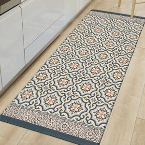 A rectangular kitchen runner rug with a geometric floral pattern in blue, cream, and orange hues on a wooden floor near white cabinetry.