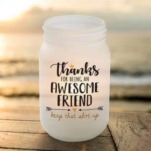 A frosted Mason jar candle displays the message, \“Thanks for being an awesome friend, keep that sh*t up,\“ in decorative fonts with crown and arrow accents, set against a blurred beach sunset background.