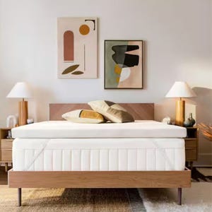 A modern bedroom setup featuring a wooden bed frame with a white mattress and several cushions. Two abstract artworks hang above the bed, flanked by matching bedside tables and lamps.