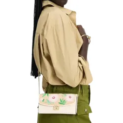A person is wearing a tan shirt and green pants, carrying a small cream shoulder bag with floral embroidery and a chain strap.