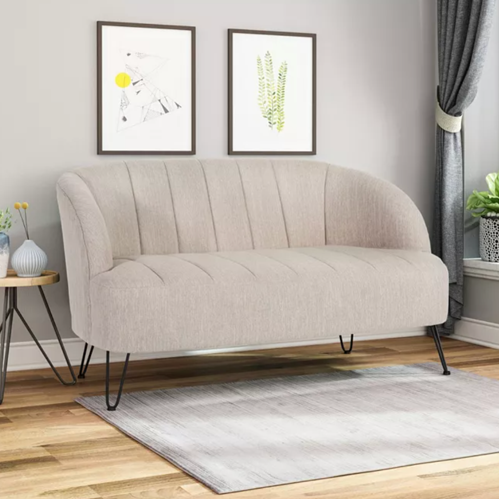 A beige upholstered loveseat with curved arms and channel tufting sits on a small gray rug alongside a round side table with vases. The room includes two framed artworks, curtains, and a potted plant on a window sill.