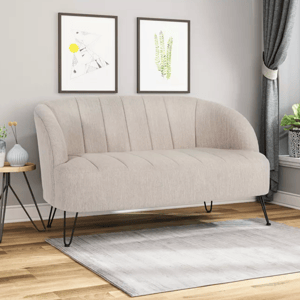 A beige upholstered loveseat with curved arms and channel tufting sits on a small gray rug alongside a round side table with vases. The room includes two framed artworks, curtains, and a potted plant on a window sill.