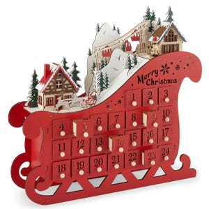 A red and white wooden advent calendar shaped like a sleigh features 24 numbered compartments and a wintry scene with houses and trees on top.