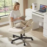 A person sits cross-legged on a beige, cushioned swivel office chair using a tablet. Nearby is a white desk with a laptop, books, and a pencil holder, set in a bright room with a large window.