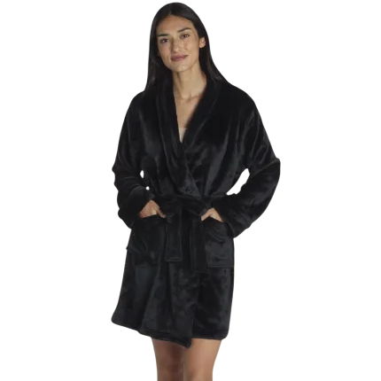 A person wearing a black, plush, knee-length bathrobe with a shawl collar, tied at the waist, standing in a modern bedroom setting.