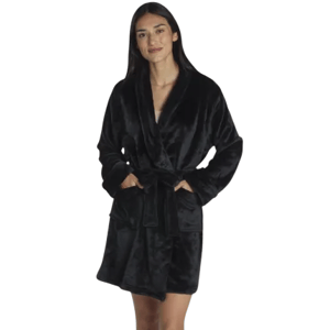 A person wearing a black, plush, knee-length bathrobe with a shawl collar, tied at the waist, standing in a modern bedroom setting.