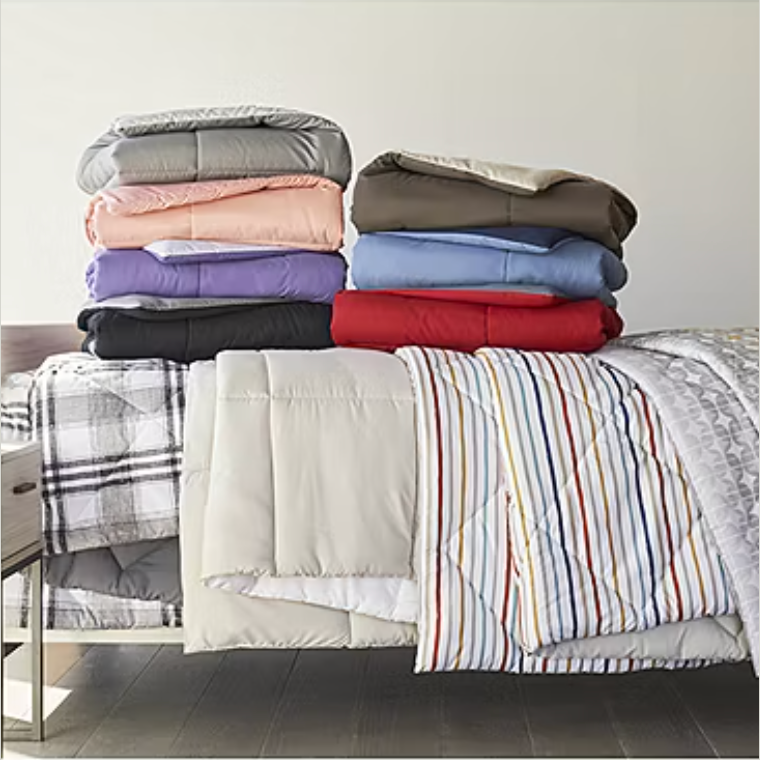 A variety of colorful, folded bed comforters are stacked on a bed along with different patterned designs.