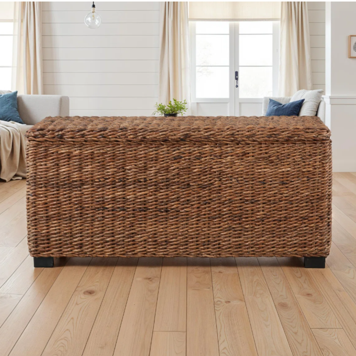A woven wicker storage bench with a natural finish is situated on a wooden floor in a living room setting, featuring a comfortable seating area in the background with neutral-colored curtains and furnishings.