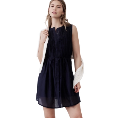 Sleeveless navy mini dress with a pleated front and a drawstring waist, paired with a white sweater draped over the shoulders.