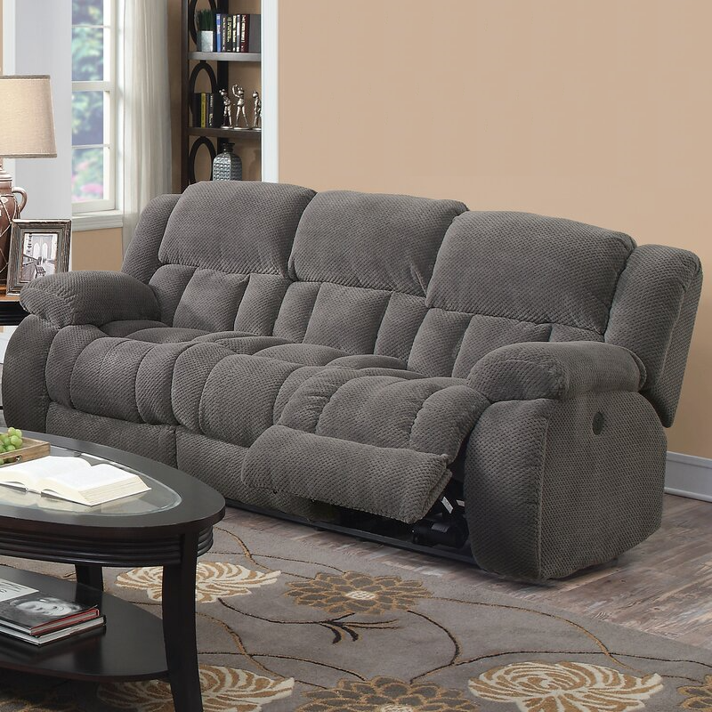 A gray reclining sofa with plush cushions is situated in a living room with a beige wall and decorative elements like a dark wood coffee table and a patterned rug.