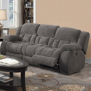 A gray reclining sofa with plush cushions is situated in a living room with a beige wall and decorative elements like a dark wood coffee table and a patterned rug.