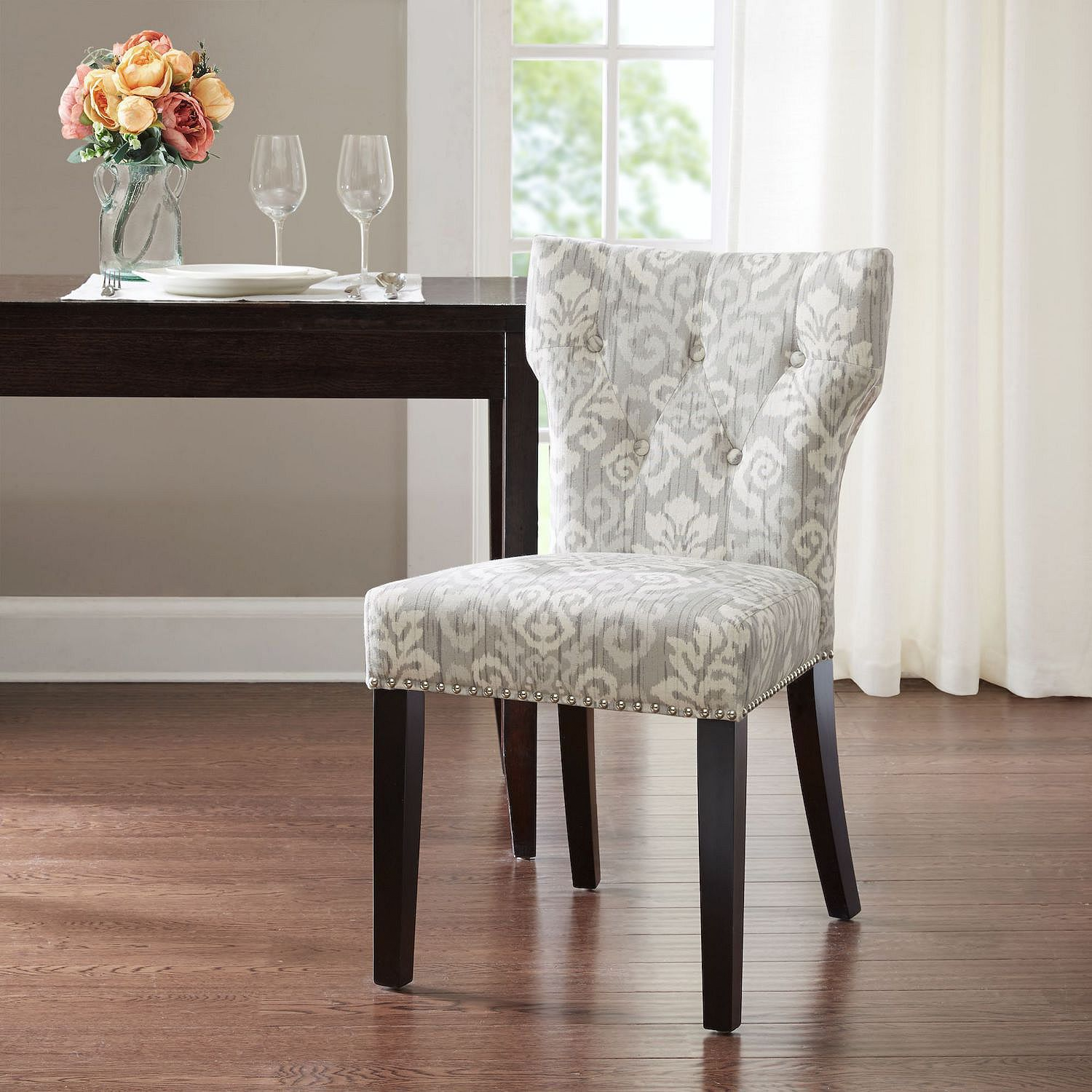 A stylish dining chair with a gray and white patterned fabric, dark wooden legs, and nailhead trim is positioned in a modern dining room setting beside a dark wooden table with two wine glasses and a vase of flowers.