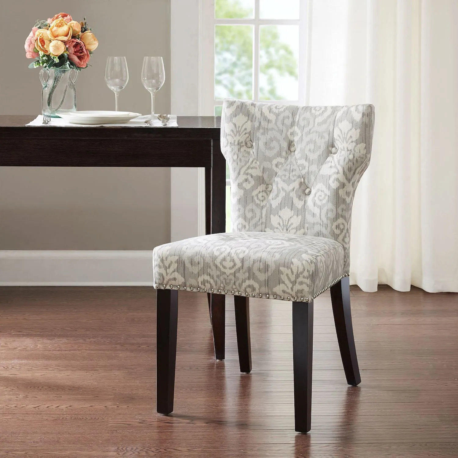 A stylish dining chair with a gray and white patterned fabric, dark wooden legs, and nailhead trim is positioned in a modern dining room setting beside a dark wooden table with two wine glasses and a vase of flowers.