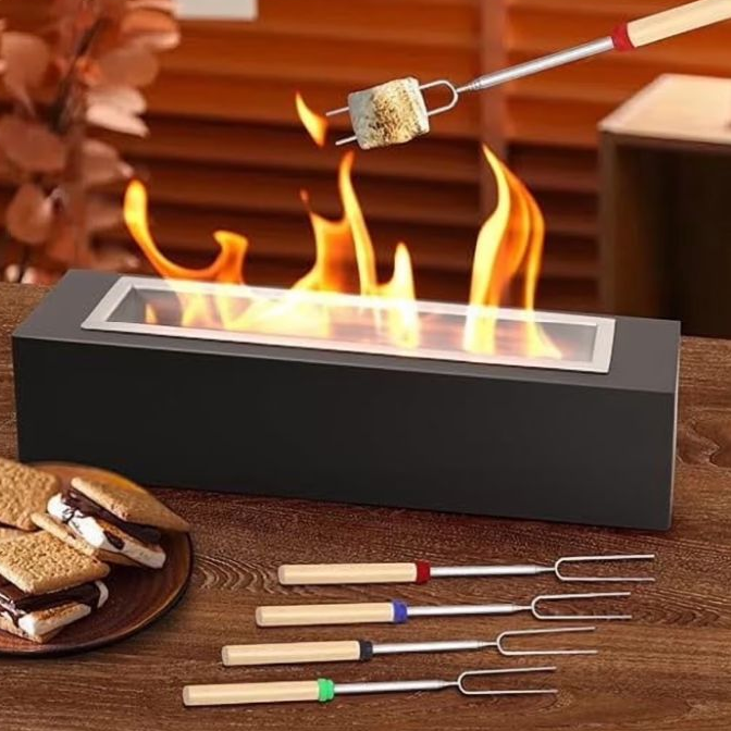 A tabletop fire pit is shown with a marshmallow being toasted on a fork. Beside it are assembled s'mores and four roasting forks with wooden handles.