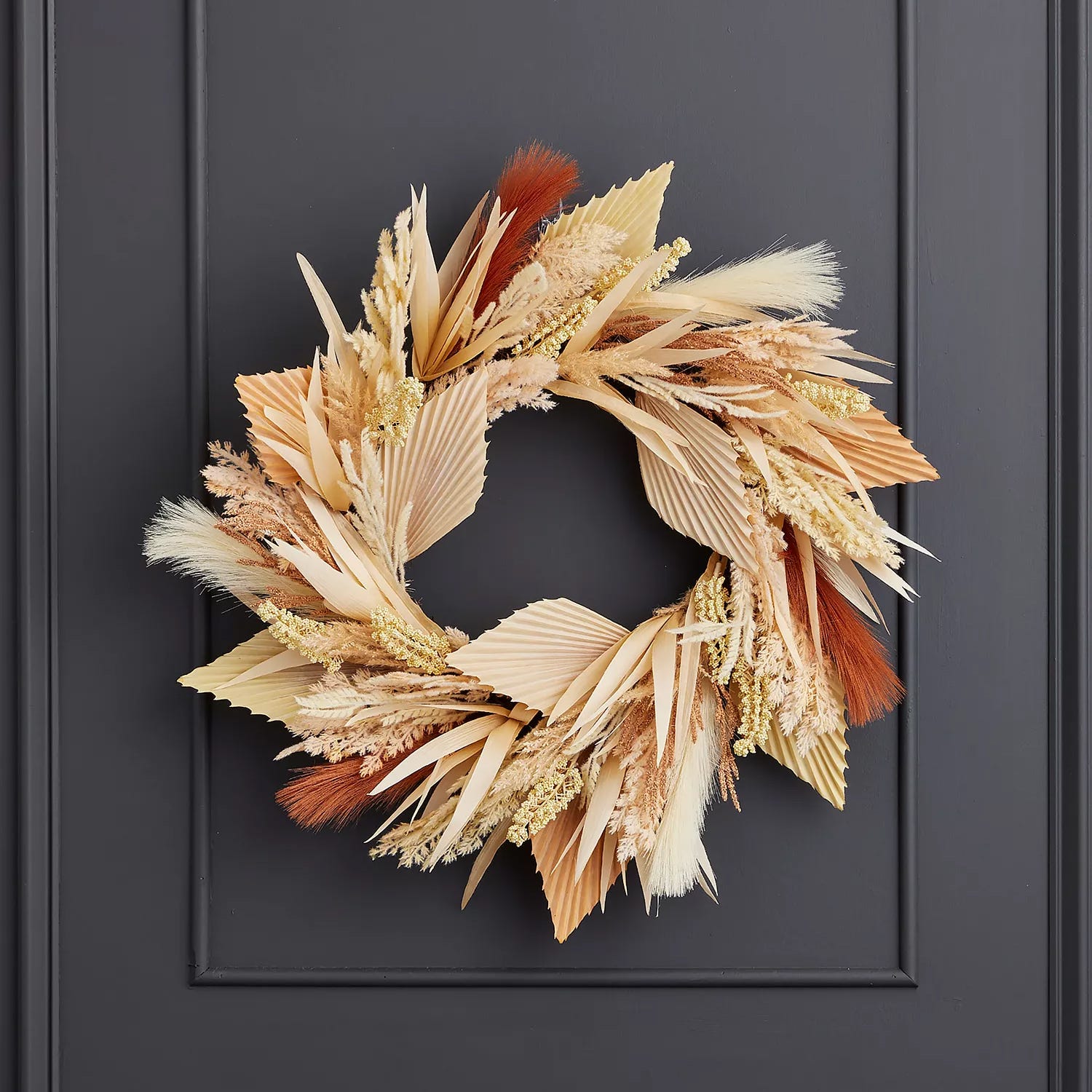 A decorative wreath featuring dried grasses and fan-shaped palm leaves in shades of beige, cream, and rust, mounted on a dark door.