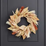 A decorative wreath featuring dried grasses and fan-shaped palm leaves in shades of beige, cream, and rust, mounted on a dark door.