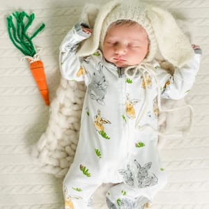 A baby is wearing Burt's Bees Baby Bunny Pajamas featuring a white base with gray and cream bunny prints and subtle green leaf accents. The outfit includes a cozy knit hat with floppy bunny ears.