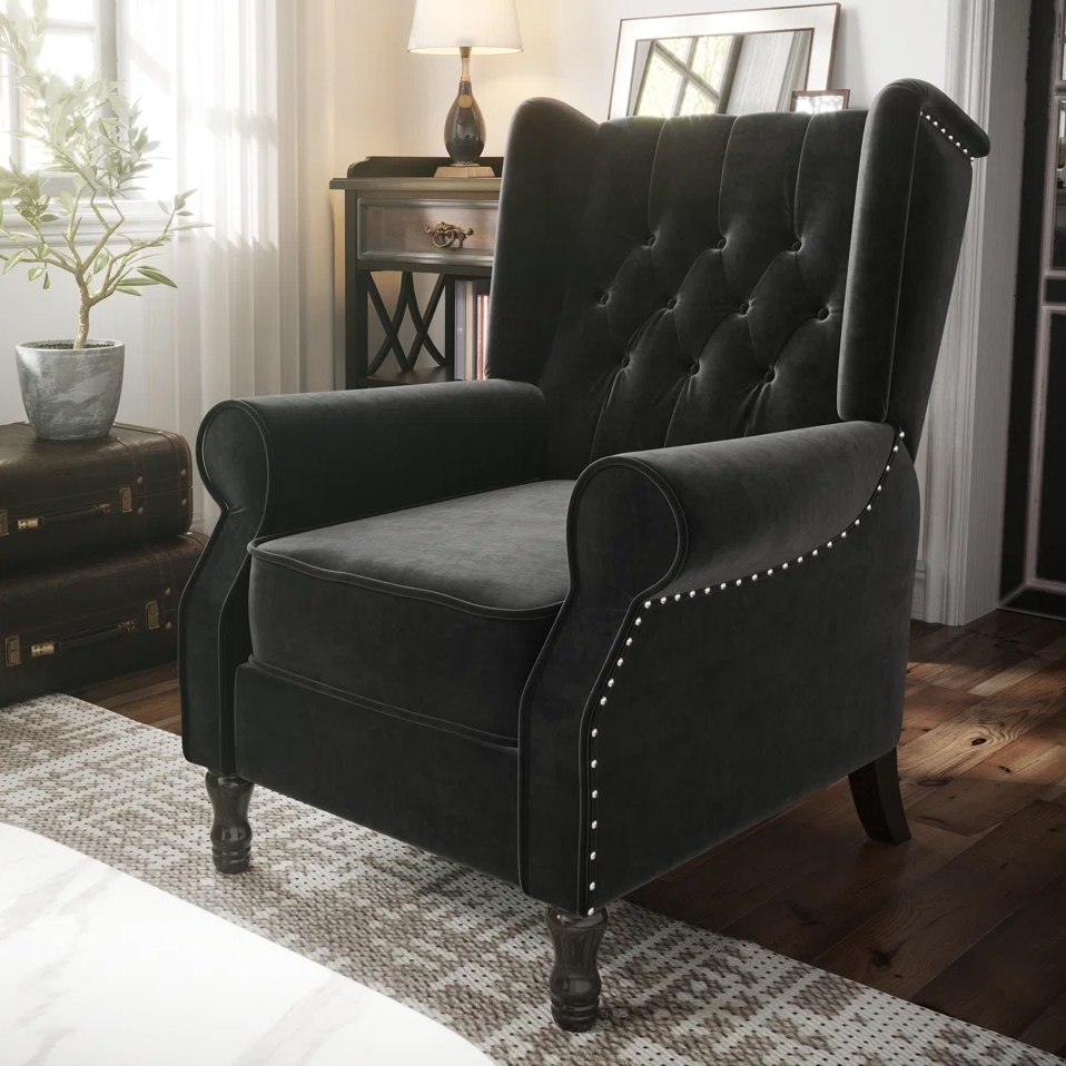 A black velvet wingback armchair with nailhead trim and button-tufted backrest, set on wooden legs. It's placed in a cozy room with a lamp, plant, and side table.