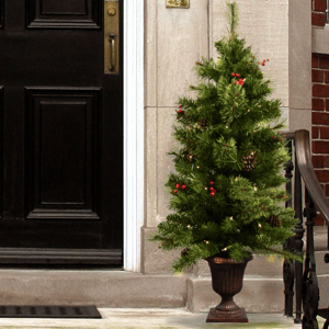 A small artificial Christmas tree in a decorative pot, adorned with lights, red berries, and pinecones, placed outside next to a black front door.