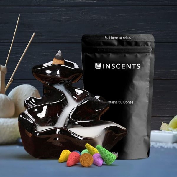 Ceramic backflow incense burner in a tiered waterfall design, accompanied by a pouch labeled \“INSCENTS\“ containing 50 cones. Various colorful incense cones are displayed in front, with a stick burner and decorative elements in the background.