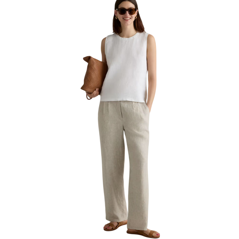 Woman in a sleeveless white top, beige wide-leg linen pants, brown sandals, and sunglasses holding a brown woven bag.
