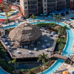 Aerial view of a tropical resort area featuring a thatched-roof pavilion with tables and chairs, surrounded by a lazy river, palm trees, and colorful seating arrangements.
