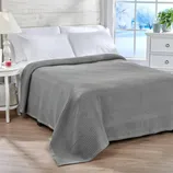 A neatly made bed with a gray waffle-textured blanket, two white pillows, and a white side table with a lamp and small items. A window shows a snowy landscape outside.