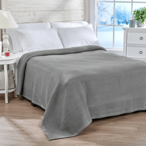 A neatly made bed with a gray waffle-textured blanket, two white pillows, and a white side table with a lamp and small items. A window shows a snowy landscape outside.